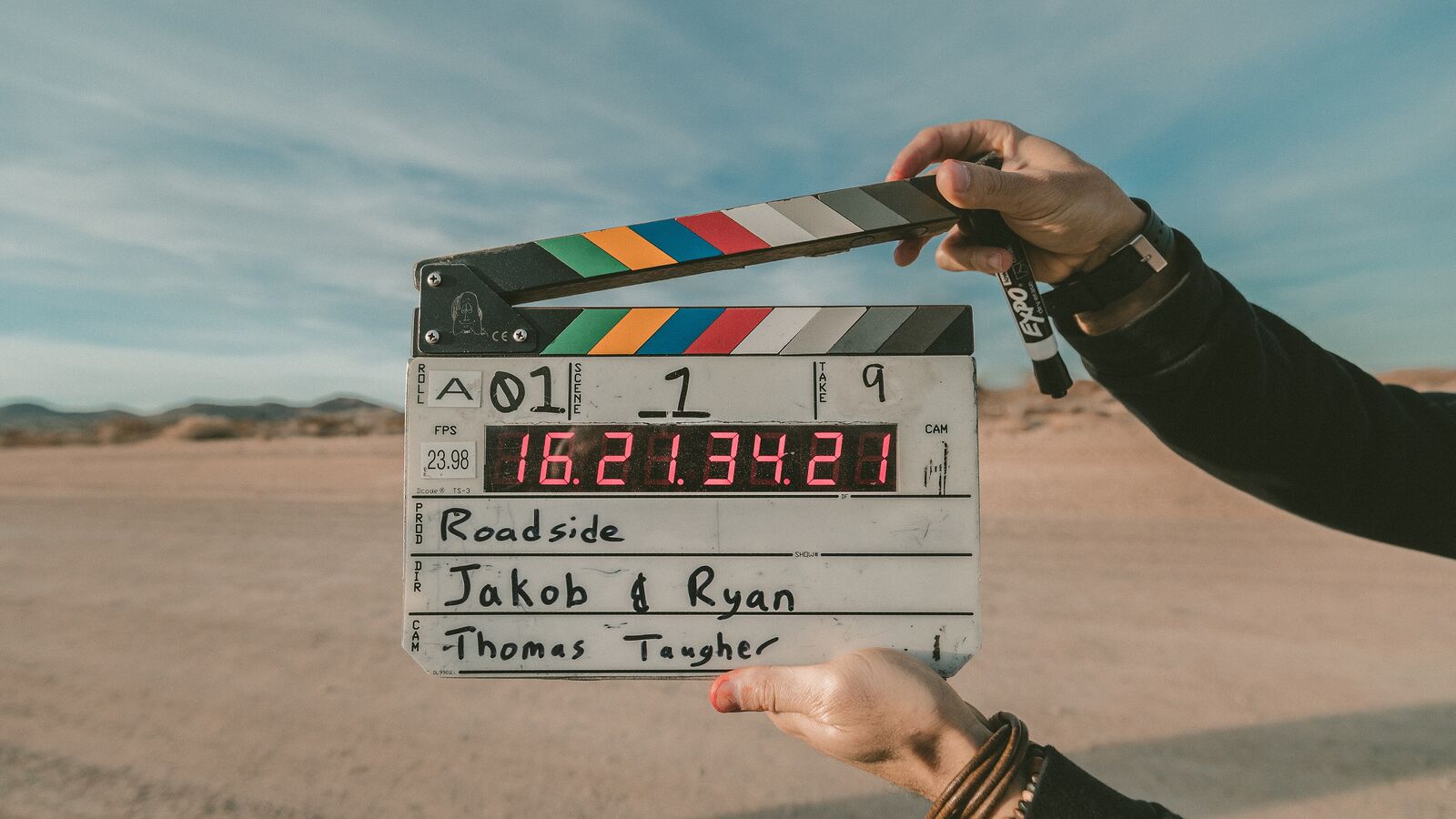 Hände halten eine geöffnete Filmklappe vor einer sandigen Landschaft und Himmel