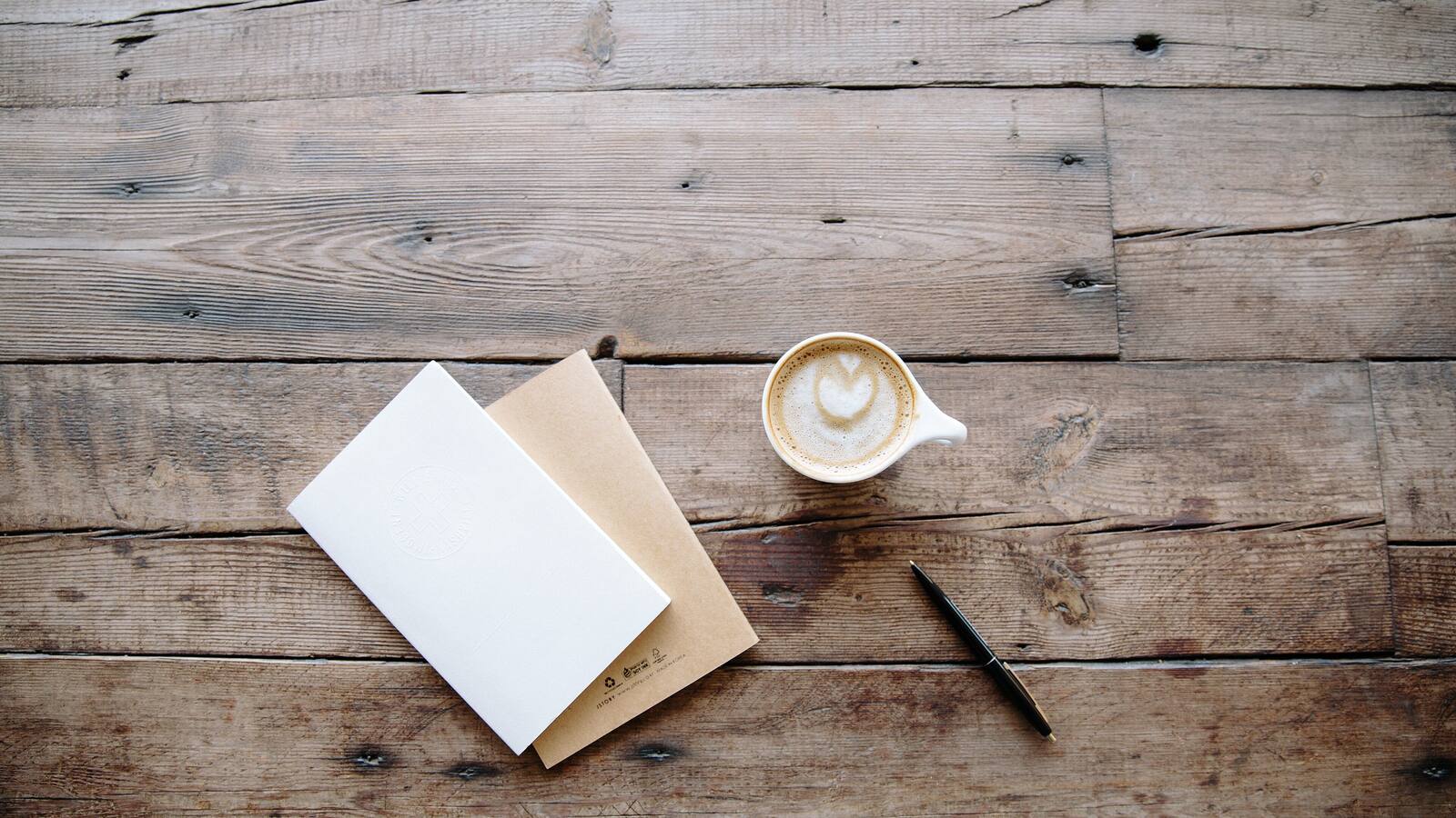 A latte, a small notebook and a pen on a rustic wooden table