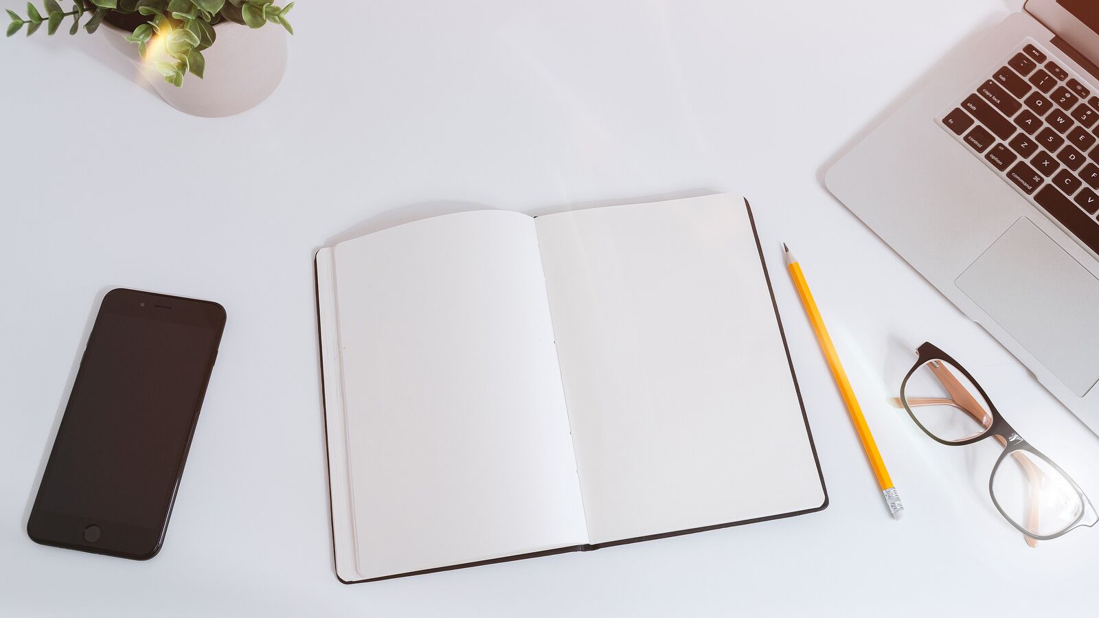 Draufsicht auf einen weißen Schreibtisch mit aufgeschlagenem leerem Notizbuch, Bleistift, Brille und Smartphone