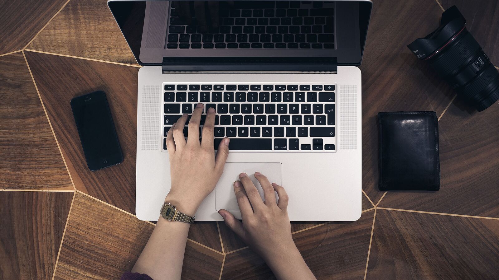 Draufsicht auf zwei Hände, die auf einem Laptop tippen, daneben ein Smartphone und ein Kameraobjektiv
