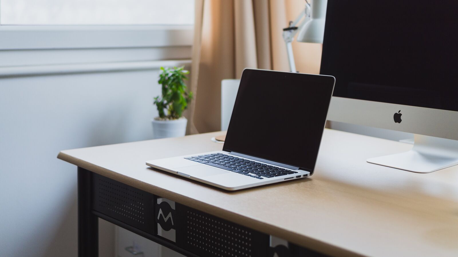 A laptop on a tidy wooden desk in a bright room with a monitor behind it